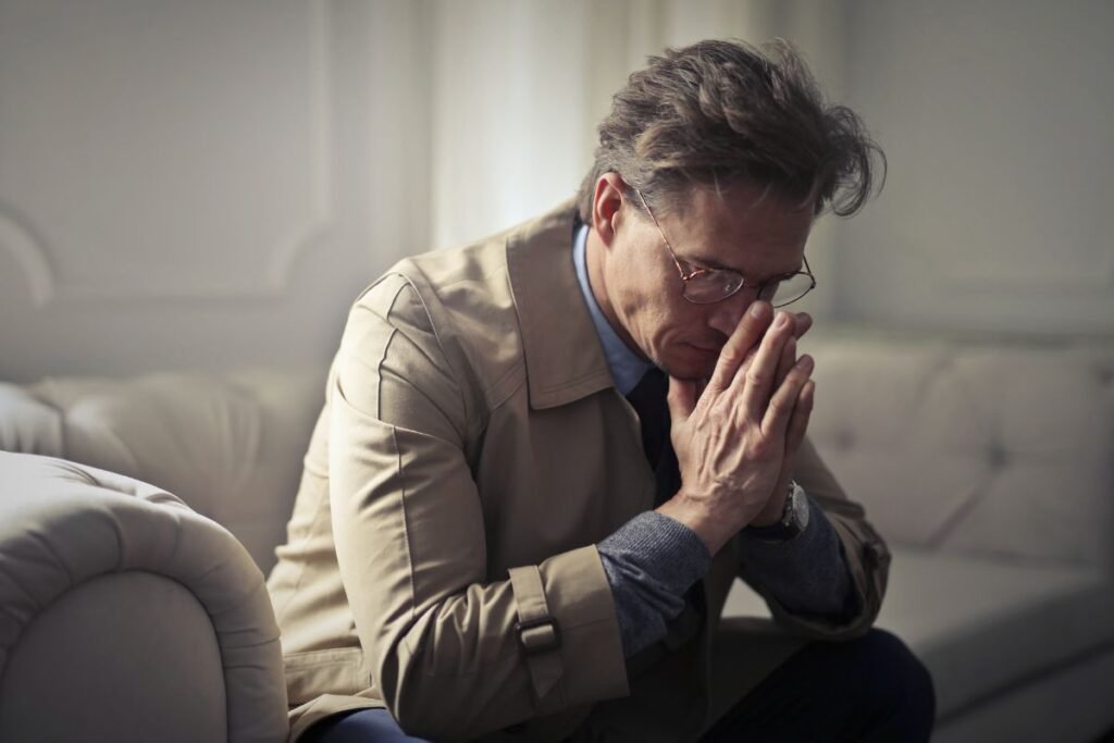 Cadre senior en pleine réflexion, assis dans son salon, les mains jointes devant son visage, illustrant la fatigue mentale et la perte de repères après une rupture professionnelle.
