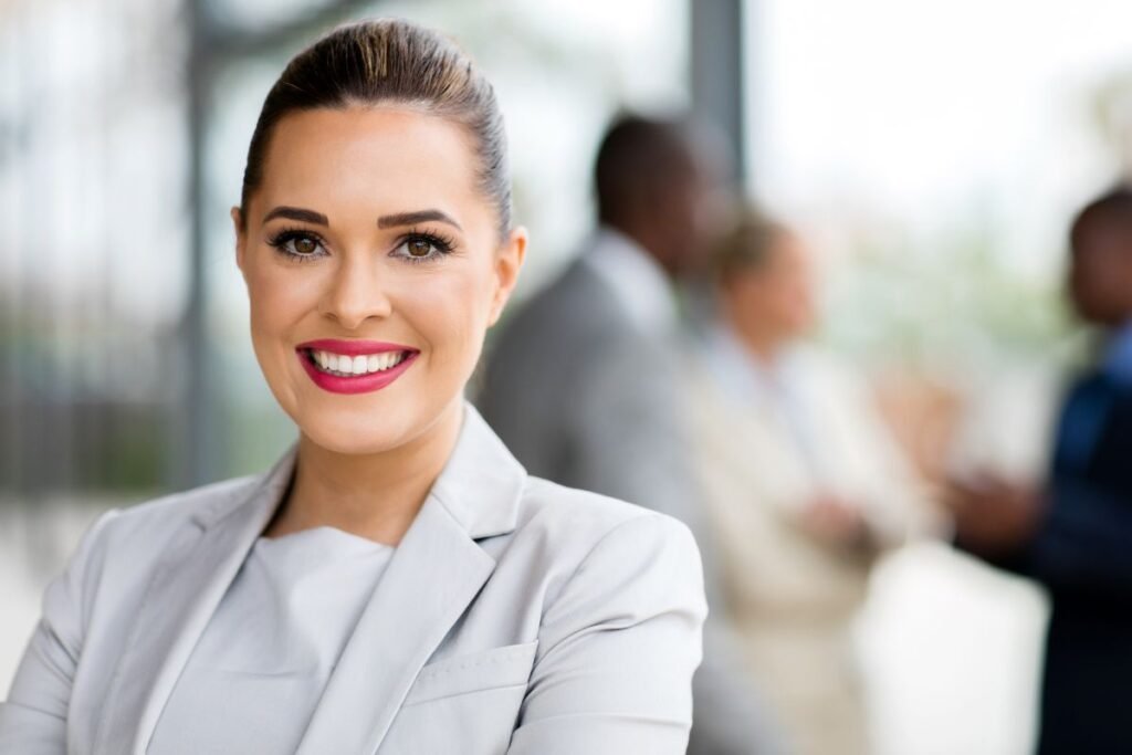 Portrait d’une femme souriante en tailleur clair, en arrière-plan flou d’un bureau avec des collègues, symbole de confiance et de sérénité professionnelle.