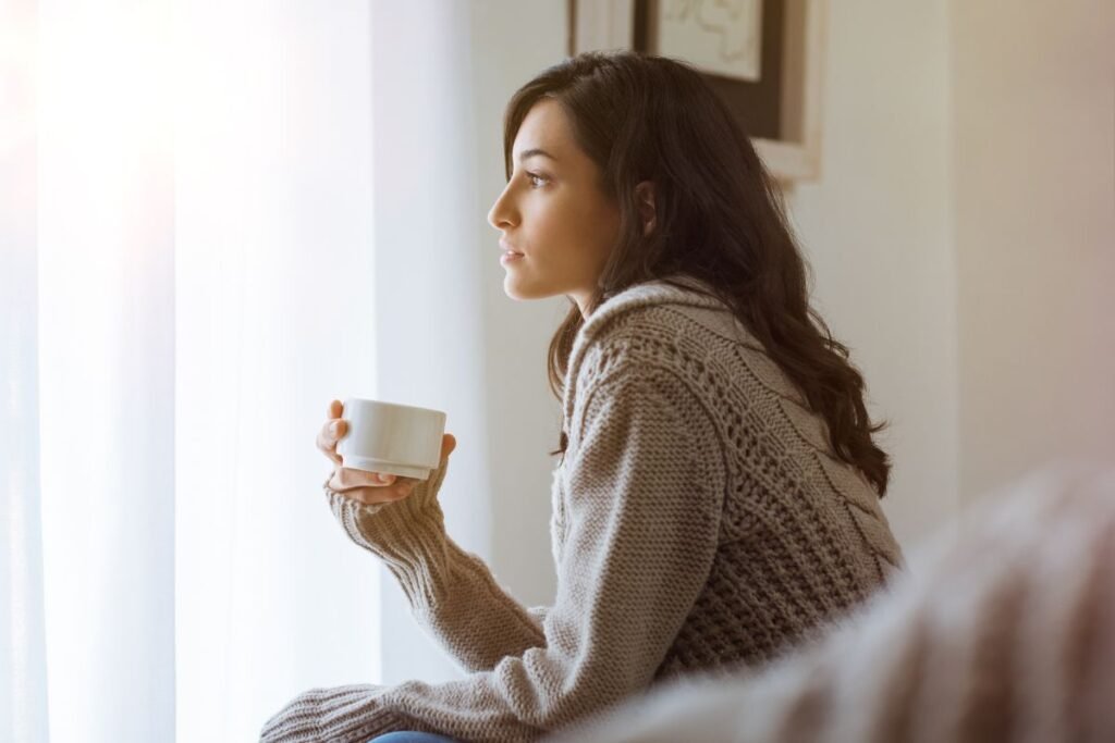 Femme en pleine réflexion chez elle, tenant une tasse de café, lumière douce du matin symbolisant un nouveau départ après une rupture professionnelle.