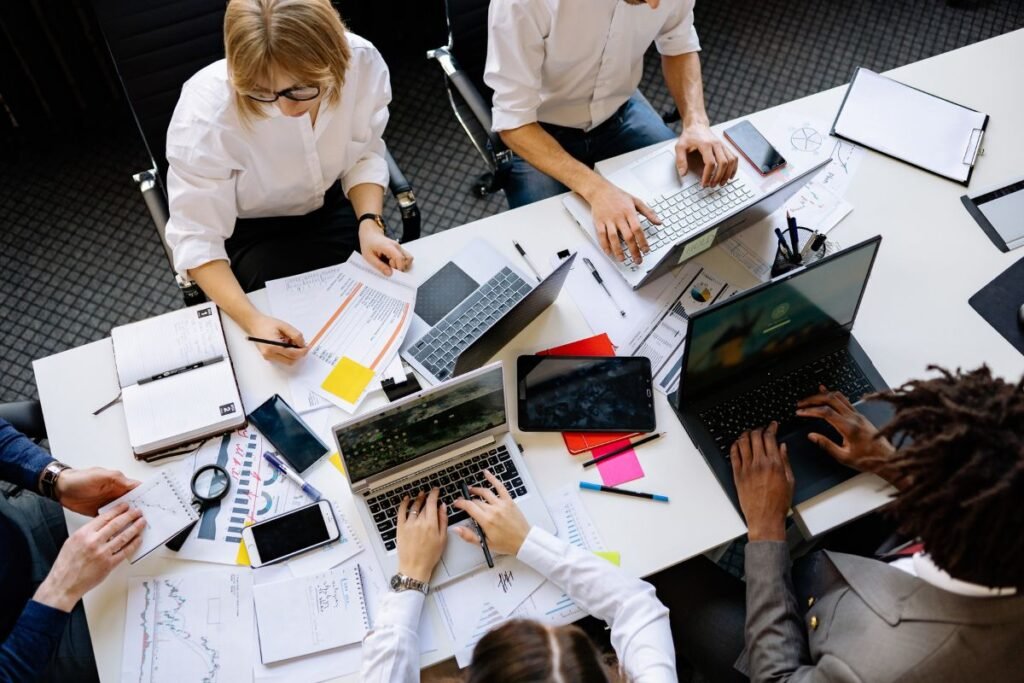 Équipe de cadres débordés en réunion, entourés de dossiers, ordinateurs et smartphones illustrant la surcharge de travail et les sollicitations multiples.