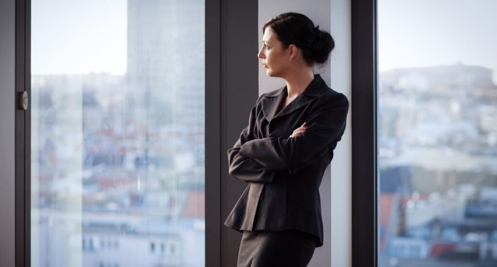 Responsable RH en costume sombre, pensive devant une grande baie vitrée, symbolisant la solitude professionnelle et la fatigue émotionnelle.