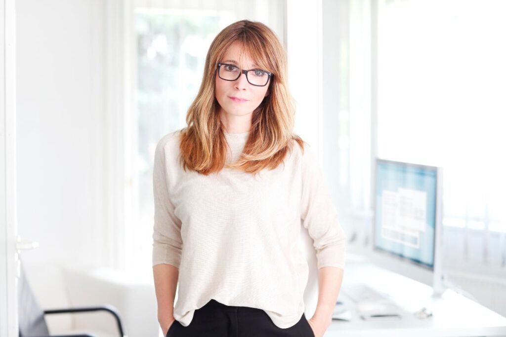Femme en blouse claire, debout dans un bureau, regard sérieux, mains dans les poches