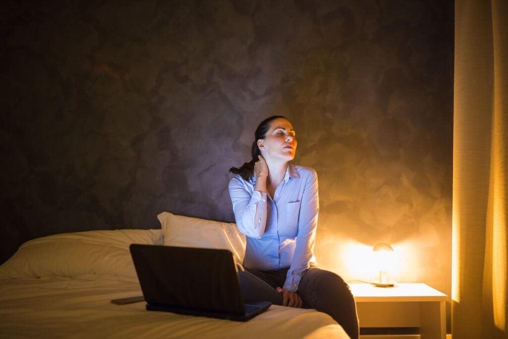 Dirigeante fatiguée assise sur son lit, tenant sa nuque douloureuse après une longue journée de travail sur ordinateur, symbole de surcharge décisionnelle et d’insomnie.