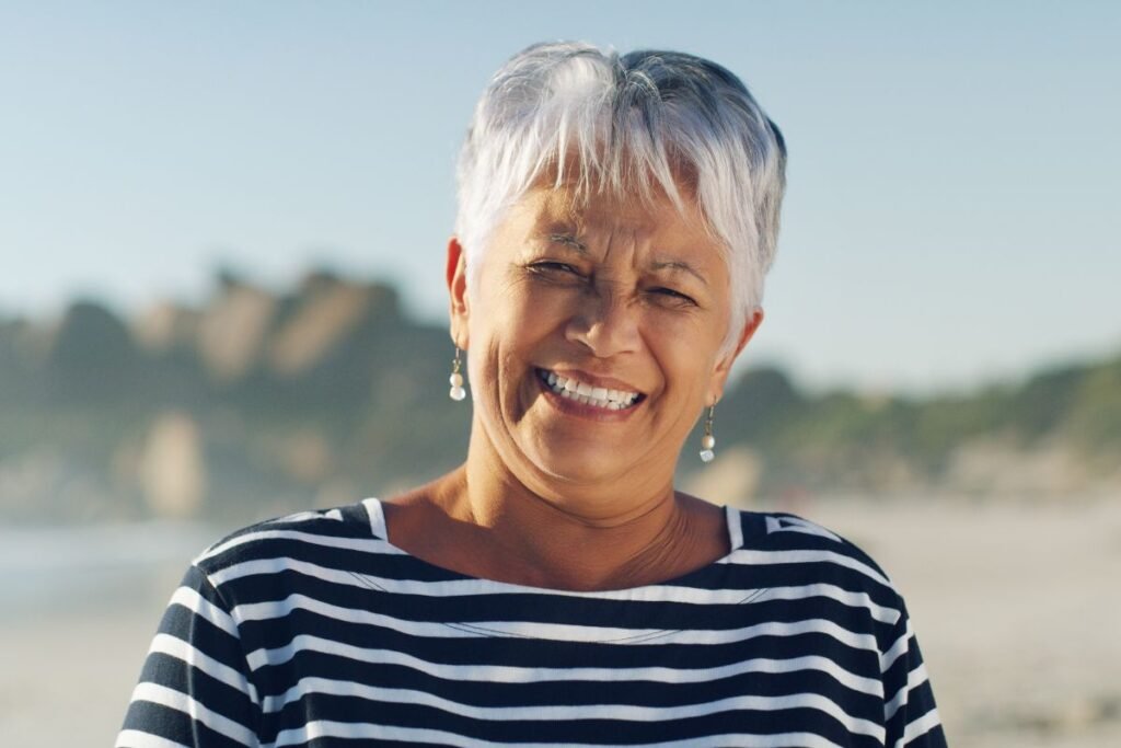 Femme souriante aux cheveux gris sur une plage, symbole de sérénité et d’épanouissement à la retraite