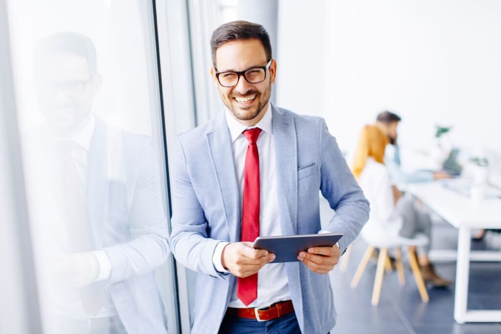 Homme en costume souriant avec une tablette, travaillant dans un environnement professionnel moderne.