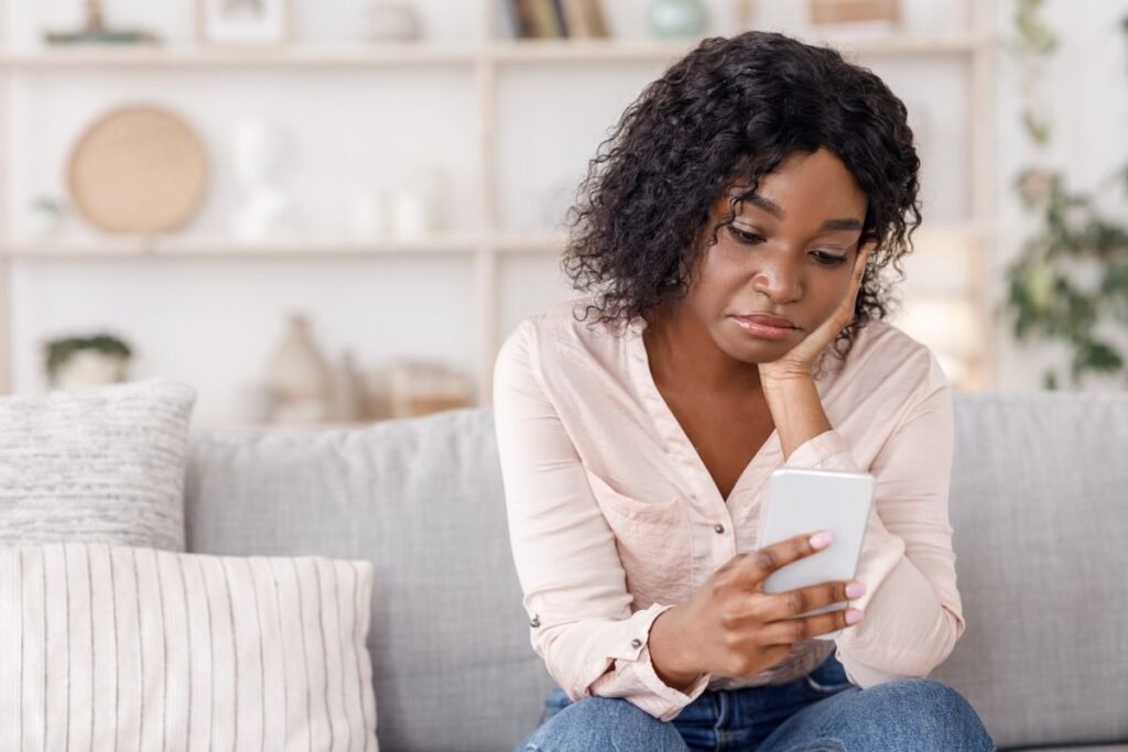Femme expatriée assise sur un canapé, regardant son téléphone avec un air pensif, illustrant la solitude à domicile.