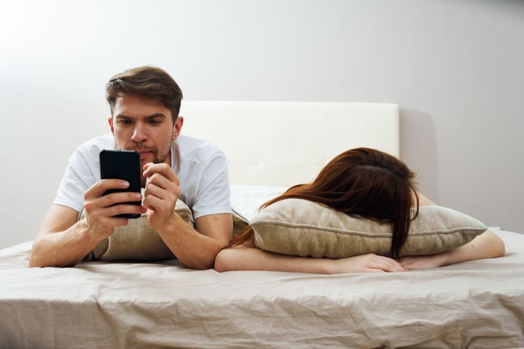 Homme absorbé par son téléphone au lit, femme allongée, illustrant les tensions conjugales liées à la charge mentale professionnelle.