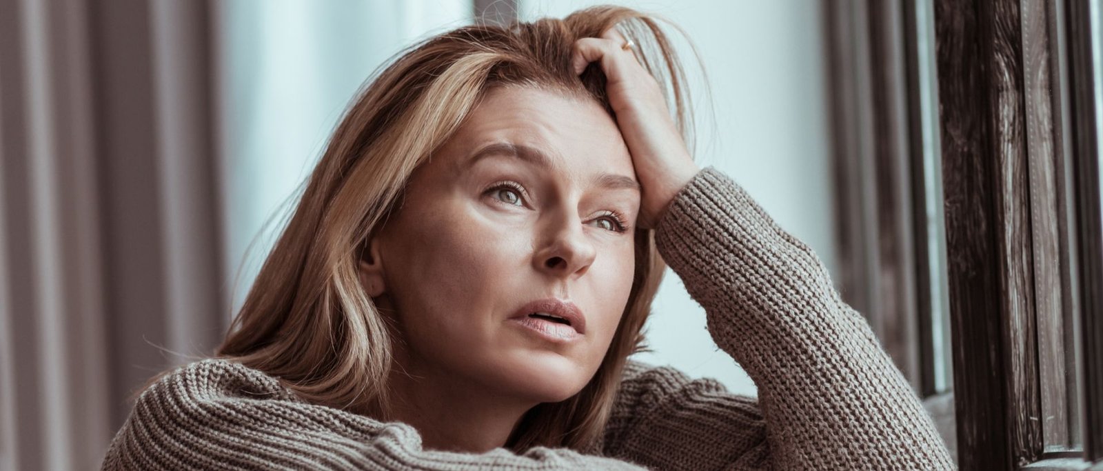 Femme expatriée pensive regardant par la fenêtre, illustrant l’éloignement des repères culturels et personnels à l’étranger.