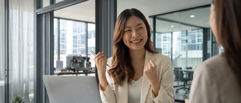 Une femme d'affaires souriante et enthousiaste, poings serrés, discute avec une collègue devant un ordinateur portable dans un bureau moderne. Cette image illustre la réussite et le sentiment de liberté après avoir appris à couper mentalement du travail.