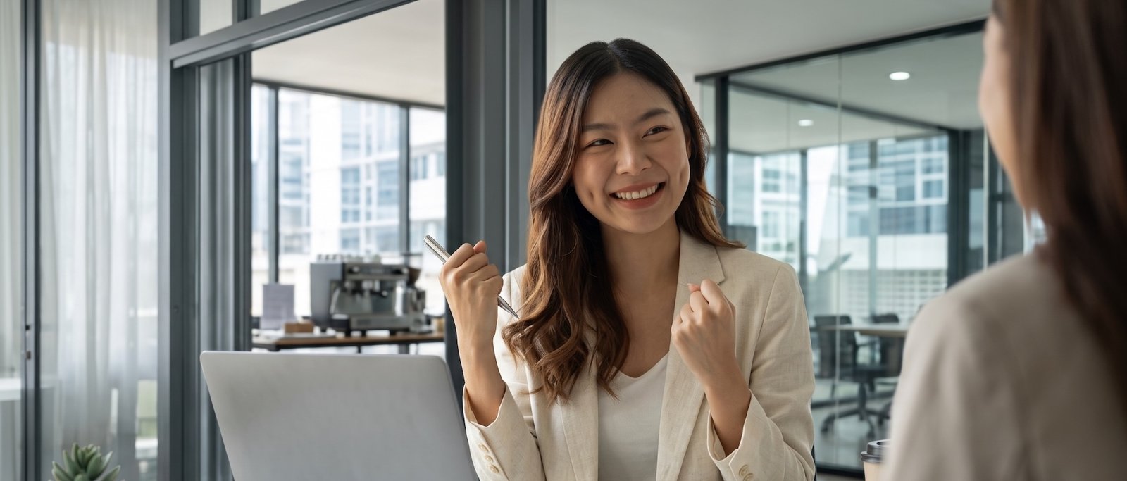 Une femme d'affaires souriante et enthousiaste, poings serrés, discute avec une collègue devant un ordinateur portable dans un bureau moderne. Cette image illustre la réussite et le sentiment de liberté après avoir appris à couper mentalement du travail.
